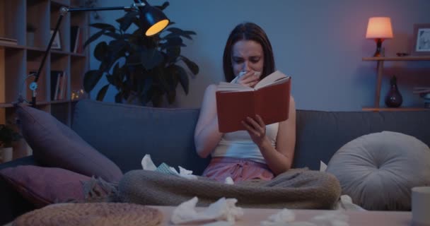 Вид спереду молодої дівчини поглинається читанням книги, сидячи на дивані. Сумна тисячолітня жінка, яка виглядає стурбованою під час читання роману та використання паперових тканин. Домашній фон — стокове відео