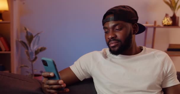 Primo piano di un giovane africano che usa il suo smartphone per chattare sui social network. Uomo barbuto indossa berretto guardando sulla dispalia telefono mentre seduto sul divano a casa. Concetto di svago . — Video Stock
