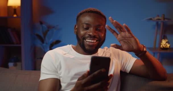 Vista frontal de cara jovem acenando e conversando ao ter uma conversa em vídeo no smartphone. Boa comunicação homem olhando usando aplicativo de telefone e sorrindo enquanto sentado no sofá em casa . — Vídeo de Stock