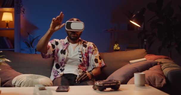 Előnézet fiatal afro férfi használ virtuális valóság headset, miközben ül a kanapén. Férfi visel vr szemüveg mozog a kezét a böngészés, görgetés, zoom, miközben otthon pihen. — Stock videók