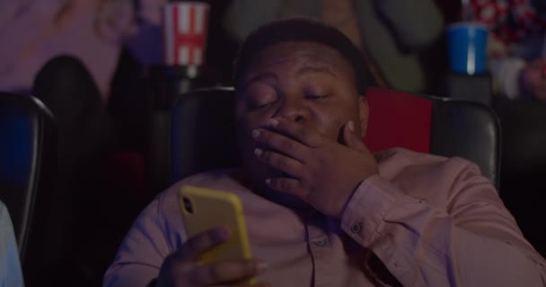 Close up of young man bored of film,using his smartphone while sitting in movie theatre. Fat millennial man yawning and covering mouth with hand while looking at his mobilephone screen. — Stock video