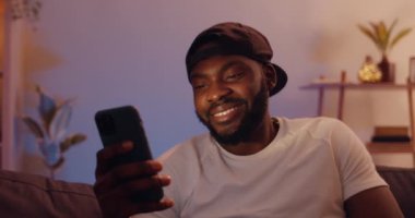 Kanepede oturmuş akıllı telefon ekranına bakarken gülümseyen neşeli genç adam. Mutlu Afrikalı adam cep telefonunu evde kullanırken iyi haberler veriyor. Boş zaman kavramı..