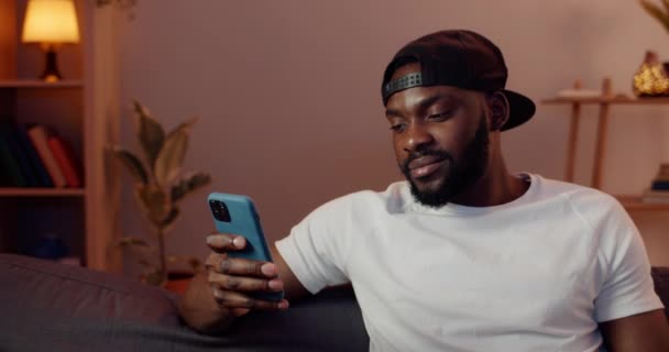 Närbild av afrikansk kille som använder sin smartphone medan du sitter på soffan hemma. Skäggig man med mössa rulla nyhetsflöde och titta på mobiltelefon skärmen medan du tillbringar ledig tid. — Stockvideo