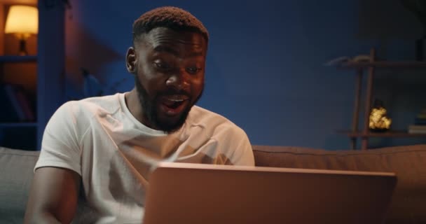 Jóképű fiatalember használja a laptopját, és boldog, amikor jó híreket kap. Szakállas afrikai srác néz személyes számítógép képernyőn, és így meglepett arc, miközben ül a kanapén. — Stock videók