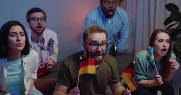 Focus on excited guy with german flags in hands rejoicing shouting and singing hymn in front of TV . Smiling man sitting near upset friends while watching sport together at home. Royalty Free Stock Video