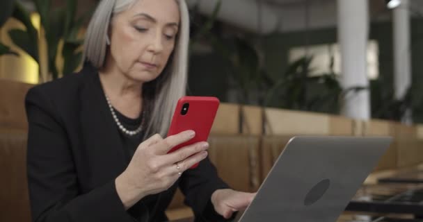 Primer plano de la anciana de pelo gris que trabaja en la cafetería con teléfono inteligente y portátil. Madura elegante mujer de negocios en auriculares utilizando sus aparatos mientras está sentado en el restaurante. En interiores . — Vídeo de stock