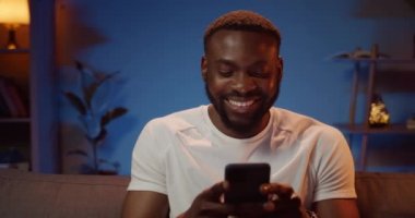 Neşeli bir Afrikalı adam akıllı telefon kullanırken arkadaşlarıyla sohbet ediyor. Oturma odasında kanepede otururken mesajlar yazan ve gülümseyen sakallı genç bir adam. Teknoloji ve insan kavramı.