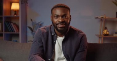 Sıradan giyinmiş, kameraya bakıp gülümseyen yakışıklı bir adam. Evdeki kanepede otururken mutlu görünen Afro-Amerikan genç adam portresi. İnsan kavramı.