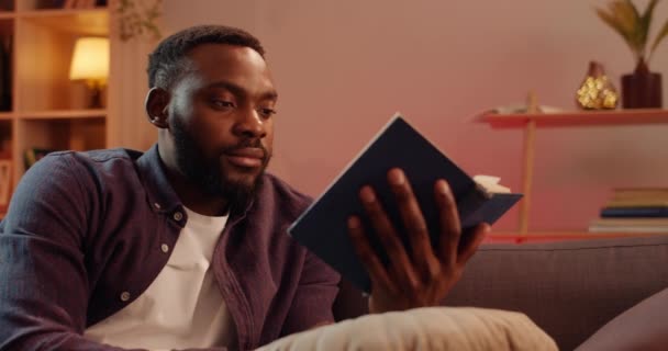 Young bearded man rubbing his eyes and looking tired of reading book. Handsome guy student reading late at night and feeling sleepy while sitting on sofa with book.Concept of leisure. — Stock Video