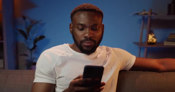 Young man in casual t shirt using smartphone for browsing net. Handsome bearded guy scrolling news feed while sitting on sofa at home. Concept of leisure and tech. — Stock Video