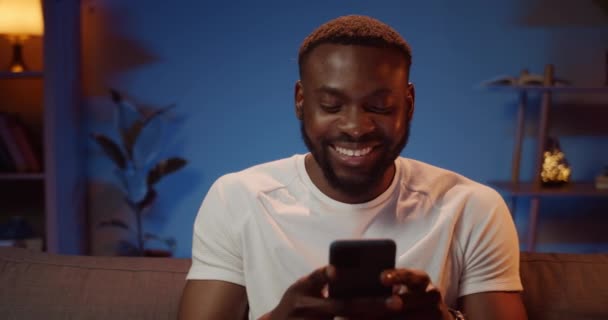 Allegro uomo africano che chatta con gli amici mentre utilizza lo smartphone. Barbuto giovane ragazzo digitando messaggi e sorridendo mentre seduto sul divano in soggiorno. Concetto di tecnologia e persone . — Video Stock