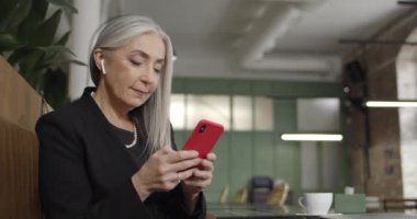 Kulaklık takan, akıllı telefon kullanan ve e-posta yazan yaşlı iş kadınının yan görüntüsü. Modern olgun, zarif bir bayan kafede oturuyor ve mesaj yazıyor. İnsan kavramı, iletişim ve teknoloji.