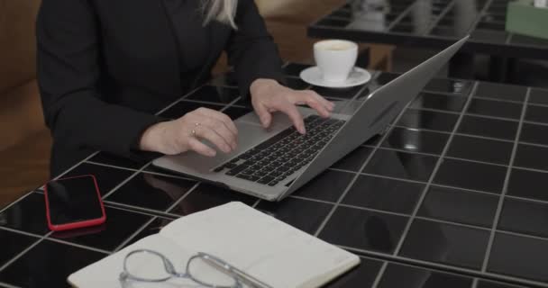 Cropped view of woman siting and working at stylish black table with coffee cup and smartphone on it. Close up of womans hands typing on laptop keyboard. Cafe background. Concept of tech. — Stock Video