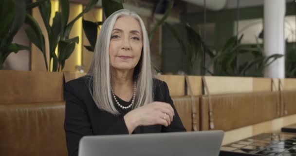 Beautiful middle aged businesswoman sitting at cafe table and working with laptop. Portrait of independent old female enterpreneur putting hand under chin and looking to camera . — Stock Video