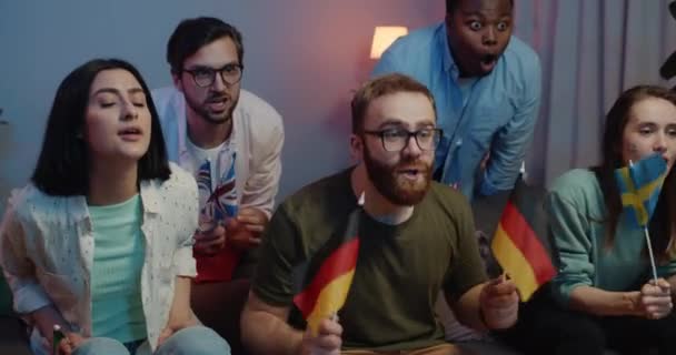 Happy caucasian man with german flags in hands rejoicing and shouting . Excited young man sitting near upset friends while watching sport game on TV at home. Concept of win and loss. — Stock Video