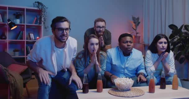 Grupo de personas alegres sentadas en el sofá y viendo deportes en la televisión en casa. Jóvenes de carrera mixta gritando, aplaudiendo y dando chocos mientras celebran el éxito de su equipo . — Vídeo de stock