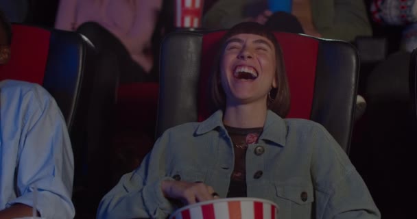 Primer plano de cara de chica sonriente viendo una divertida película de comedia en el cine. Mujer milenaria sentada y riendo en el cine mientras come palomitas de maíz. Concepto de entretenimiento . — Vídeo de stock