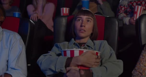 Крупный план реакции девушки на страшный момент в кино. Молодая хипстерша смотрит ужастик в кинотеатре и обнимает попкорн. Концепция развлечений и досуга — стоковое видео