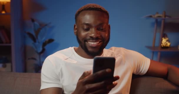Jeune homme souriant utilisant une caméra frontale sur son smartphone pour discuter avec un ami. Heureux gars assis sur le canapé, parlant et gesticulant tout en ayant un appel vidéo. Accueil arrière-plan . — Video
