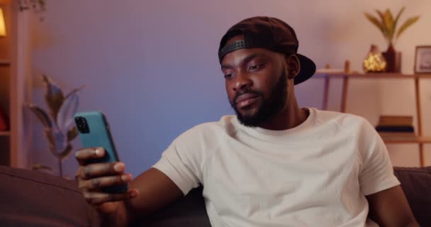 Relaxed young man using his mobile phone for browsing net while spending time at home. Handsome guy wearing cap sitting on sofa and looking at smartphone screen. Concept of leisure. — Stock Video