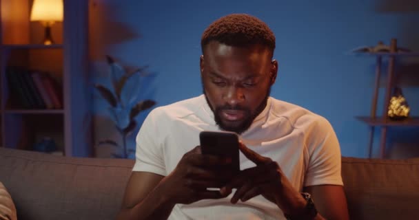 Handsome young guy using his smartphone and looking happy when getting good news. Bearded african man holding mobile phone in hands and making surprised face while sitting on sofa. — Stock Video