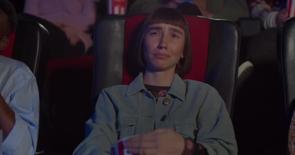 Młoda kobieta ogląda smutny dramat w kinie i przytula popcorn bowl. Hipsterka płacze i wyciera łzy ręką, patrząc na ekran w kinie. Pojęcie rozrywki . — Wideo stockowe
