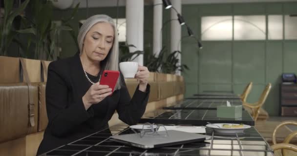 Madura elegante mujer de negocios sentada en la mesa de la cafetería durante el descanso. Emprendedora anciana usando smartphone y bebiendo cofee. En el interior. Concepto de empresa, trabajo y personas . — Vídeos de Stock