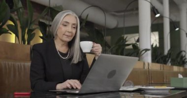 Elinde kahve fincanı olan ve dizüstü bilgisayarda çalışan olgun stil sahibi bir iş kadını. Yaşlı zarif bayan girişimci kafe masasında oturuyor ve çalışıyor. Kapalı alanda. İş kavramı.