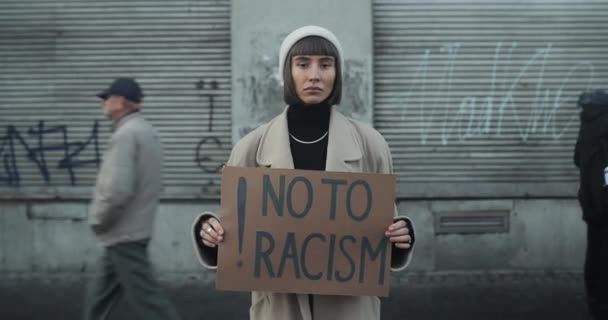 Lviv, Ukraine - November 27, 2019: Νεαρή γυναίκα που κρατά όχι στη φράση ρατσισμός χαρτόνι και στέκεται στο δρόμο. Κορίτσι που υποστηρίζει την εκστρατεία κατά του ρατσισμού. Χρονικό διάστημα. Οι άνθρωποι περπατούν στο παρασκήνιο. — Αρχείο Βίντεο
