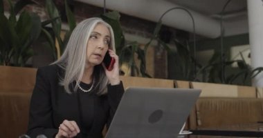 Kafede otururken akıllı telefondan konuşan konsantre kadın işyeri sahibi. Başarılı, olgun girişimci cep telefonuyla iş konuşurken dizüstü bilgisayarla çalışıyor..
