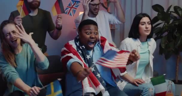 Alegre joven con bandera americana en el hombro celebrando la victoria del equipo que apoyaba. Emocionado chico afroamericano sentado cerca de amigos molestos mientras ve el deporte en la televisión . — Vídeos de Stock