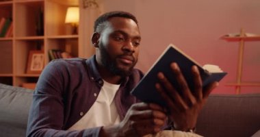 Yakışıklı Afrikalı adamın kitabı kapayıp başını sallayışını yakından çek. Genç adam oturma odasında kanepede otururken okumayı bitiriyor. Boş zaman ve hobi kavramı.