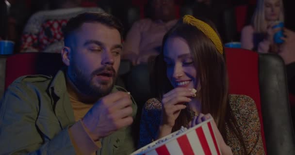 Close up of couple eating popcorn and feeding each other while watching movie in cinema. Young man and woman having good time while having date in cinema. Concept of relationship. — Stock Video