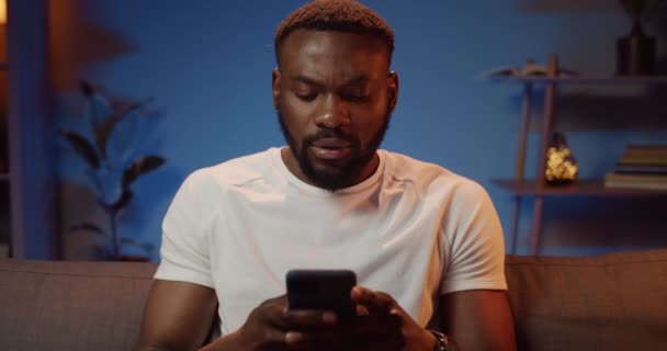 Vue de face de l'homme africain barbu en t-shirt blanc tapant message tout en étant assis sur le canapé. Beau gars utilisant le téléphone pour la communication dans les réseaux sociaux tout en passant du temps à la maison . — Video