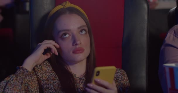 Attractive girl rolling eyes with boredom while watching a terrible film. Close up of young woman bored of film started using her smartphone in a movie theatre. Concept of entertainment. — Stock Video