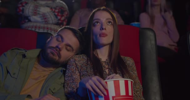 Un bel homme barbu dormant sur l'épaule de sa copine pendant un film inintéressant. Jeune jolie fille mangeant du pop-corn et regardant un film pendant que son petit ami dort. Concept de loisir . — Video