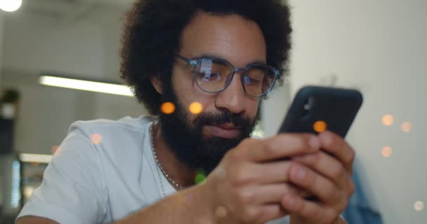 Beau homme en lunettes regardant l'écran du smartphone tout en tapant le message. Vue rapprochée du barbu dans les années 30 à l'aide de son téléphone portable pour la communication tout en étant assis. À l'intérieur . — Video