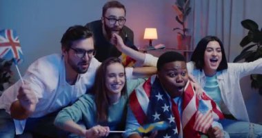 Bir grup çok ırklı arkadaş birlikte eğleniyor, TV 'de spor maçı izlerken kucaklaşıyorlar. Bayrakları olan mutlu genç insanlar, desteklerken ve tezahürat ederken birlikte kanepede oturuyorlar..