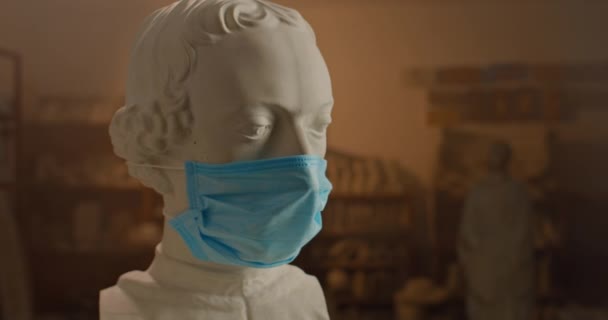Γκρο πλαν άποψη της παλιάς αντίκα λευκό μαρμάρινο προτομή γλυπτική φορώντας ιατρική μάσκα. Concept of art on qurantine.Εργαστήριο γλυπτών στο παρασκήνιο. — Αρχείο Βίντεο