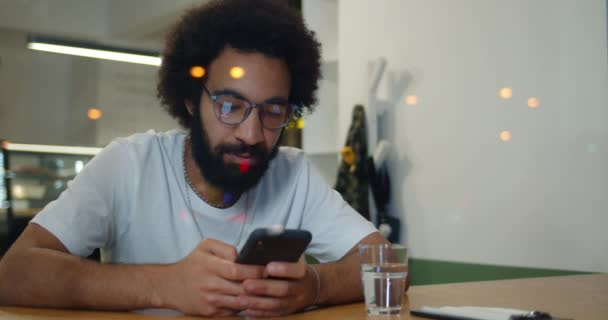Bearded guy in 30s using his smartphone and smiling while sitting in cafe. Cheerful man in glasses and t shirt chatting in social networks on phone while spending time outdoors. — Stock Video