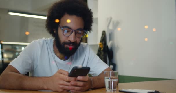 Ragazzo barbuto sulla trentina usando il suo smartphone e sorridendo mentre era seduto nel caffè. Uomo allegro in occhiali e t shirt chatta nei social network al telefono mentre trascorre del tempo all'aperto . — Video Stock