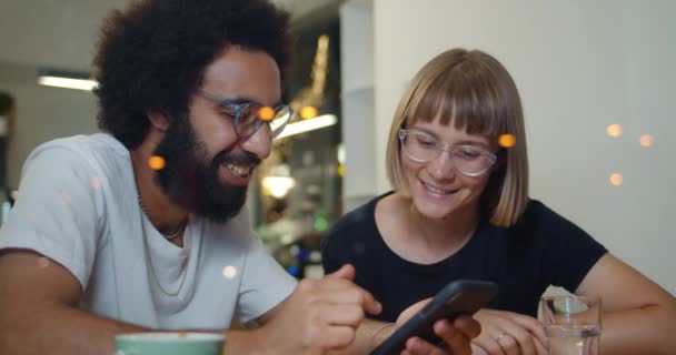 Két izgatott barát, akik jól érzik magukat együtt, miközben a modern kávézóban ülnek. Jóképű férfi és nő beszél és mosolyog, miközben képeket néz az okostelefonon. A szabadidő fogalma. — Stock videók