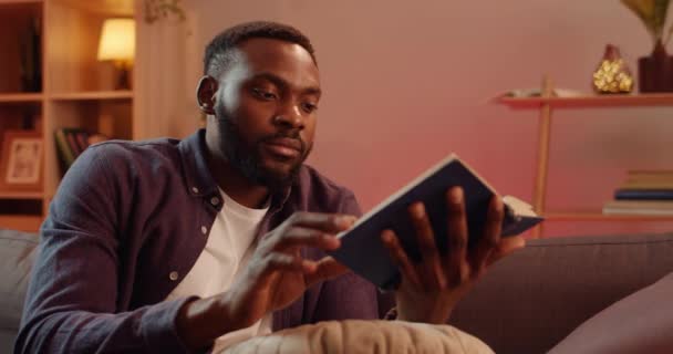 Närbild av stilig afrikansk man hålla bok medan du sitter på soffan i vardagsrummet. Ung skäggig kille i skjortöppningsbok och börjar läsa. Begreppet fritid och hobby. — Stockvideo