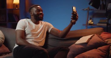 Rahat genç adam kanepede otururken ve telefon kullanırken evde vakit geçiriyor. Tişörtlü sakallı adam video sohbeti yaparken konuşuyor ve gülümsüyor. Ev arkaplanı.