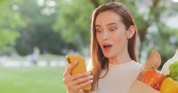 Visão de perto da mulher milenar dizendo uau e fazendo rosto surpreso ao usar o smartphone. Menina chocada bonita segurando comida no saco de papel e olhando para a tela do telefone. Ao ar livre . — Vídeo de Stock