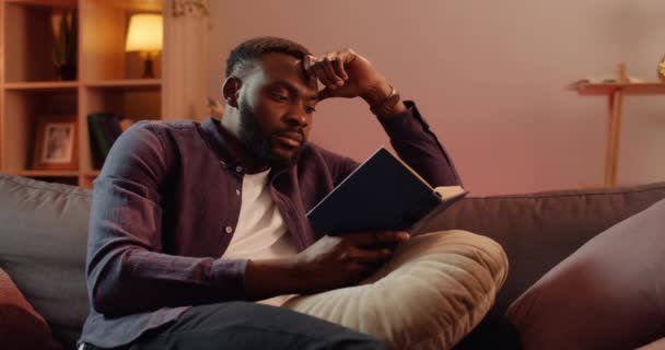 Красивый африканский парень наклоняет голову рукой, сидя на диване в гостиной. Молодой человек проводит свободное время, читая интересную книгу. Концепция досуга и хобби . — стоковое видео