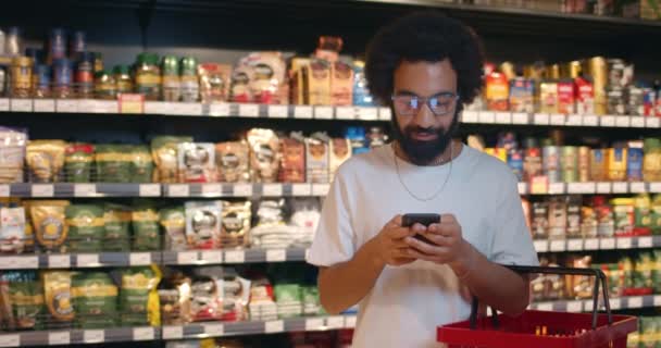 Hombre barbudo guapo en gafas usando smartphone mientras hace compras con cesta. chico maduro en los años 30 lista de verificación en el teléfono móvil, mientras que de pie en la sección de tienda de alimentos . — Vídeo de stock