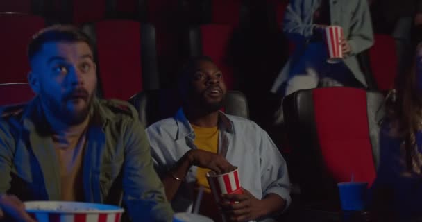 Excited spactators starting hold seats while film already started. Young african eating popcorn and watching film while other people taking their seats. Concept of leisure and entertainment. — Stock Video