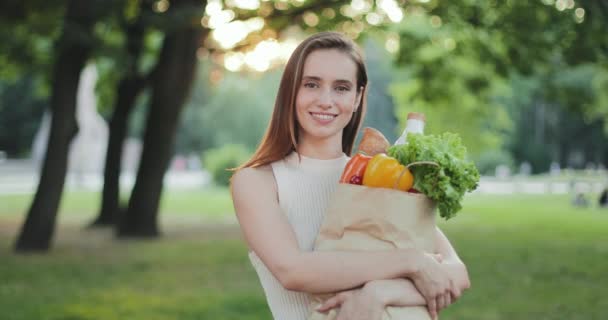 Portrait of cheerful girl looking to camera while holding paper bag with food.Beautiful young woman smiling and posing while standing with shopping. Concept of healthy lifestyle.Zoom in. — Stock Video