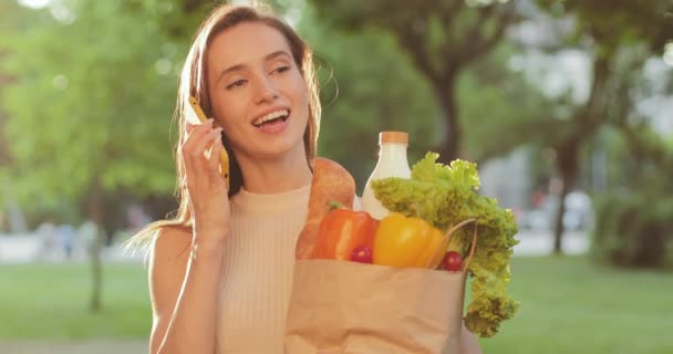 Schöne Millennial-Frau hält Papiertüte mit Lebensmitteln in der Hand und benutzt ihr Smartphone zur Kommunikation. Lächelndes Mädchen, das telefoniert und ihre Einkäufe hält, während sie draußen steht. Vergrößern. — Stockvideo